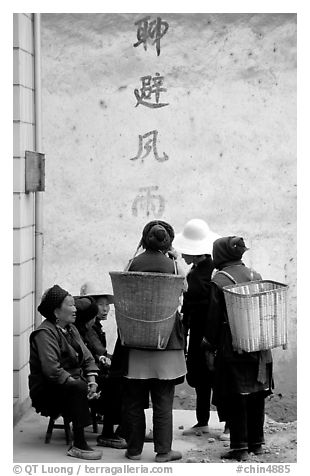 Elderly women with back baskets in front of a wall with Chinese scripture. Shaping, Yunnan, China (black and white)