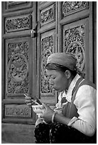 Bai woman eating from a bowl in front of carved wooden doors. Dali, Yunnan, China (black and white)