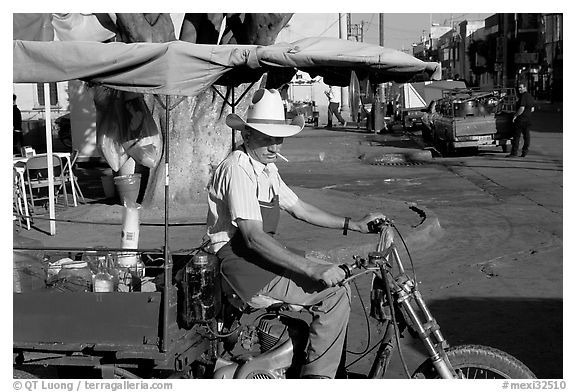 Man with cigarette riding a motorcycle-powered food stand on town plaza. Mexico