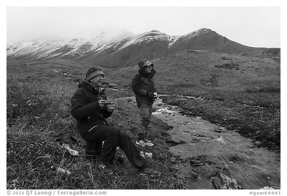 Backpackers eating by a creek and snowy mountains. Gates of the Arctic National Park (black and white)