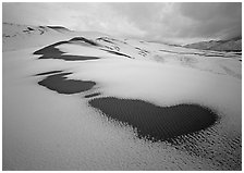 Patches of uncovered sand in snow-covered dunes, mountains, and dark clouds. Great Sand Dunes National Park, Colorado, USA. (black and white)
