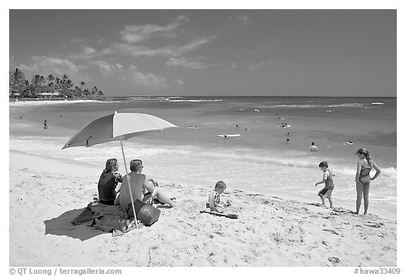 Couple sitting under sun unbrella with children playing around, Poipu Beach, mid-day. Kauai island, Hawaii, USA (black and white)