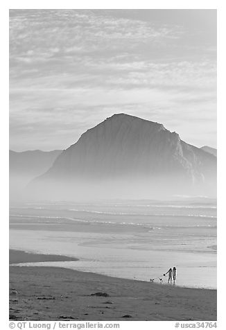 Women walking dog on the beach, with Morro Rock behind. Morro Bay, USA (black and white)