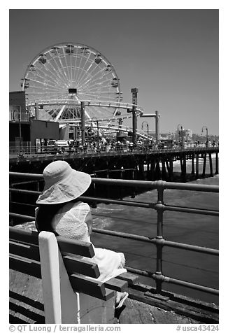 Woman sitting on bench with pink hat and ferris wheel. Santa Monica, Los Angeles, California, USA (black and white)