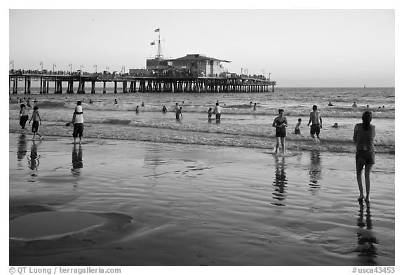 Beachgoers near Santa Monica Pier reflected in wet sand, sunset. Santa Monica, Los Angeles, California, USA (black and white)