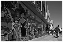 Man riding bicycle on sidewalk past mural, Mission District. San Francisco, California, USA (black and white)