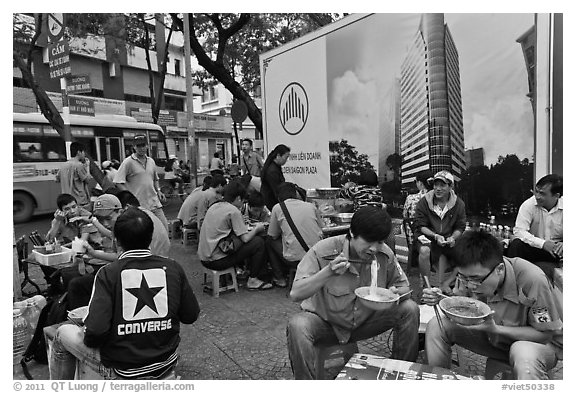 Uniformed students eating breakfast in front of backdrop depicting high rise in construction. Ho Chi Minh City, Vietnam (black and white)