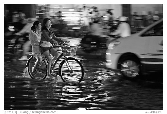 Women sharing a bicycle ride at night on a water-filled street. Ho Chi Minh City, Vietnam