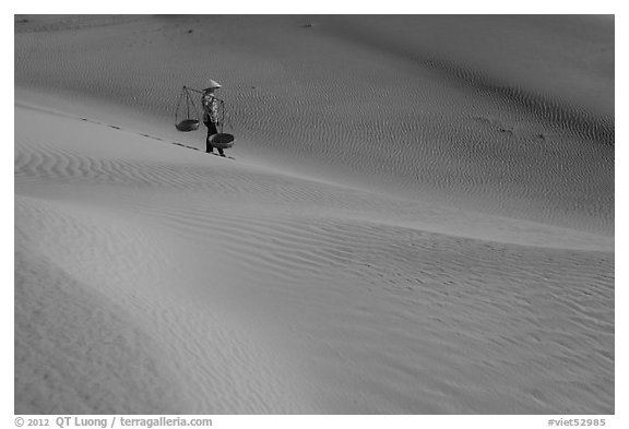 Woman with conical hat and yoke baskets pauses on sand dunes. Mui Ne, Vietnam (black and white)