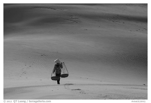 Woman walking on dune field with yoke baskets. Mui Ne, Vietnam (black and white)