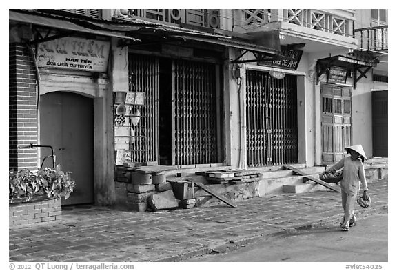 Woman carrying fruit in front of old storefronts. Hoi An, Vietnam (black and white)