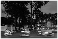 Blurred motorbikes at dusk and tall trees next to Van Hoa Park. Ho Chi Minh City, Vietnam (black and white)