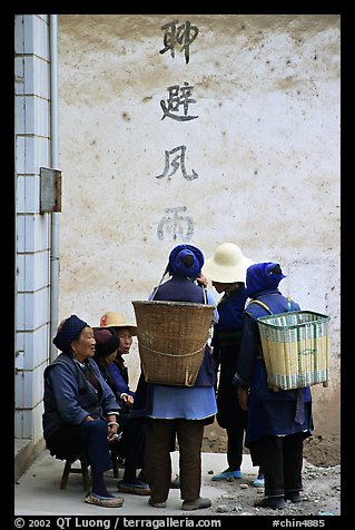 Elderly women with back baskets in front of a wall with Chinese scripture. Shaping, Yunnan, China (color)
