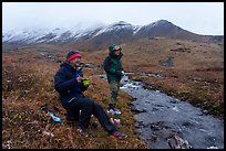Backpackers eating by a creek and snowy mountains. Gates of the Arctic National Park ( color)
