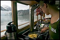 Woman prepares breakfast eggs aboard small tour boat, with glacier in view. Glacier Bay National Park, Alaska, USA. (color)