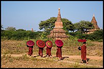 Buddhist Novices with red sun umbrellas on path near old stupas. Bagan, Myanmar