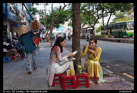Women elegantly dressed in ao dai eating on the street. Ho Chi Minh City, Vietnam