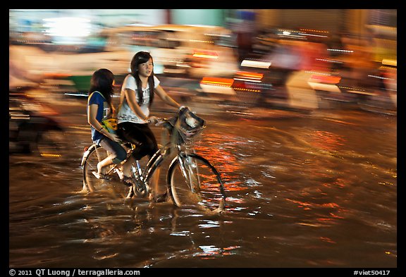 Girls sharing night bicycle ride through water of flooded street. Ho Chi Minh City, Vietnam