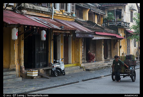 Man pulling cart in front of old townhouses. Hoi An, Vietnam (color)