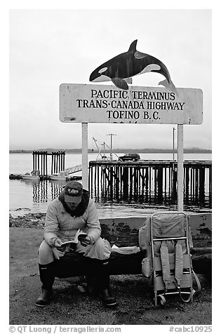 Backpacker sitting under the Transcanadian terminus sign, Tofino. Vancouver Island, British Columbia, Canada (black and white)