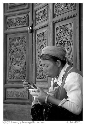 Bai woman eating from a bowl in front of carved wooden doors. Dali, Yunnan, China (black and white)