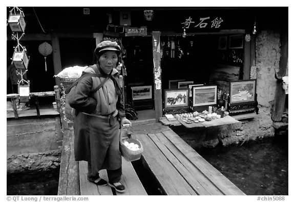 Naxi woman peddling eggs  to local residents walks acros a canal. Lijiang, Yunnan, China