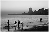 People standing in water at sunset with skyline behind, Chowpatty Beach. Mumbai, Maharashtra, India (black and white)