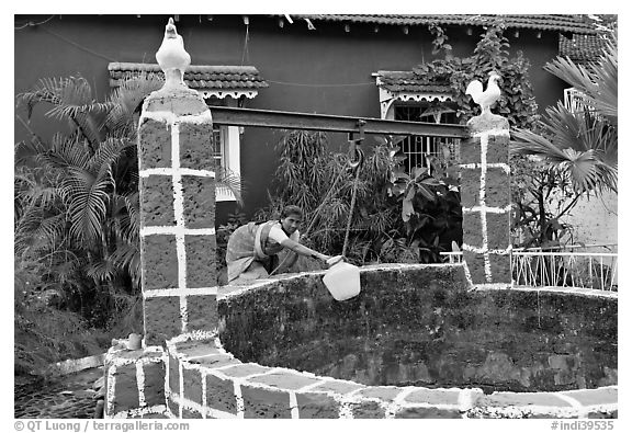 Woman retrieving water from well with blue house behind, Panjim. Goa, India (black and white)