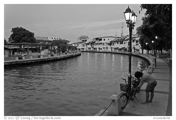 Woman locking bicyle on quay of Melaka River. Malacca City, Malaysia (black and white)