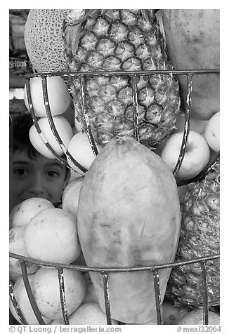 Boy peers from behind fruits offered at a juice stand, Tlaquepaque. Jalisco, Mexico (black and white)