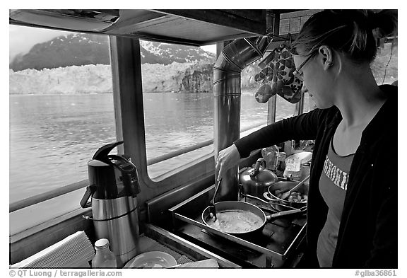 Woman cooking eggs aboard small tour boat, with glacier in view. Glacier Bay National Park, Alaska, USA.