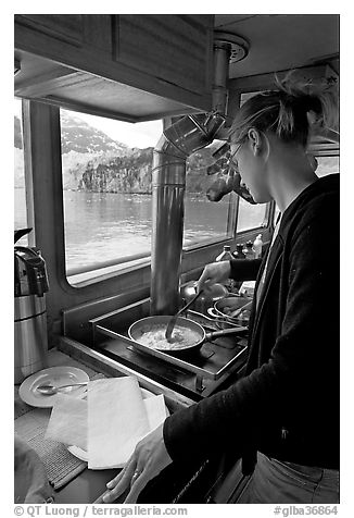 Woman cooking eggs aboard small tour boat, with glacier outside. Glacier Bay National Park (black and white)