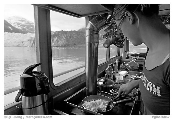 Woman prepares breakfast eggs aboard small tour boat, with glacier in view. Glacier Bay National Park (black and white)