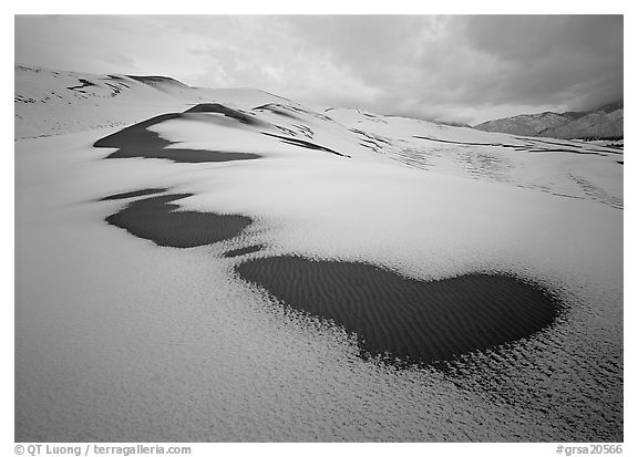 Patches of uncovered sand in snow-covered dunes, mountains, and dark clouds. Great Sand Dunes National Park, Colorado, USA.