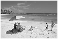 Couple sitting under sun unbrella with children playing around, Poipu Beach, mid-day. Kauai island, Hawaii, USA ( black and white)