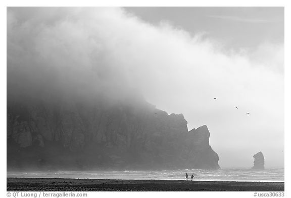 Two people strolling on the beach at the base of Morro Rock. Morro Bay, USA (black and white)