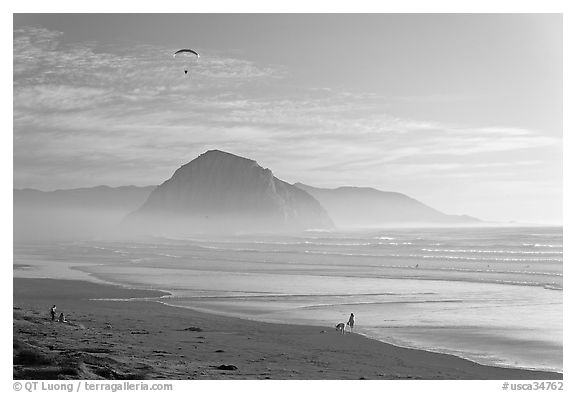 Motorized paraglider, women walking dog, with Morro Rock in the distance. Morro Bay, USA (black and white)