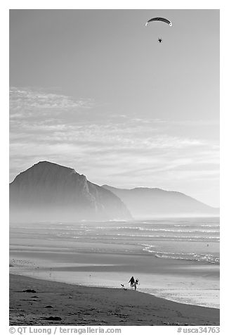 Motorized paraglider, women walking dog, and Morro Rock seen from Cayucos Beach. Morro Bay, USA (black and white)