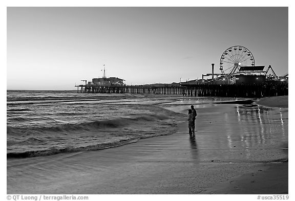 Couple reflected in wet sand at sunset, with pier and Ferris Wheel behind. Santa Monica, Los Angeles, California, USA (black and white)