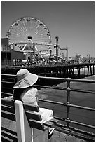 Woman sitting on bench with pink hat and ferris wheel. Santa Monica, Los Angeles, California, USA (black and white)