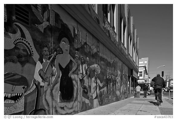 Man riding bicycle on sidewalk past mural, Mission District. San Francisco, California, USA (black and white)