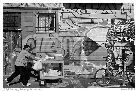 Man pushes vending cart pass mural and bicycle, Mission District. San Francisco, California, USA (black and white)