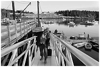 Man carrying construction wood and rolling case out of mailboat. Isle Au Haut, Maine, USA (black and white)