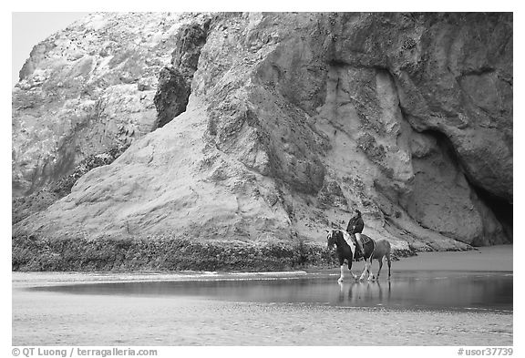 Woman horse-riding on beach next to sea cave entrance. Bandon, Oregon, USA (black and white)