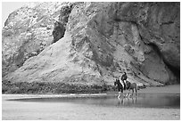 Woman horse-riding on beach next to sea cave entrance. Bandon, Oregon, USA (black and white)