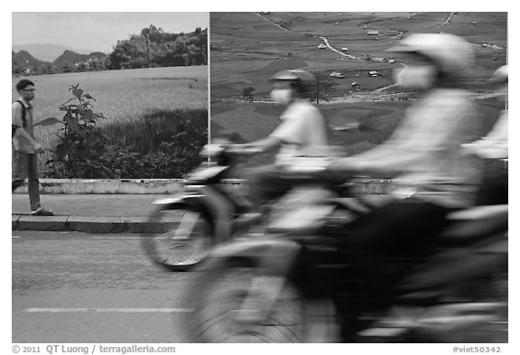 Man walking and motorbike riders blured in front of backdrops depicting traditional landscapes. Ho Chi Minh City, Vietnam (black and white)
