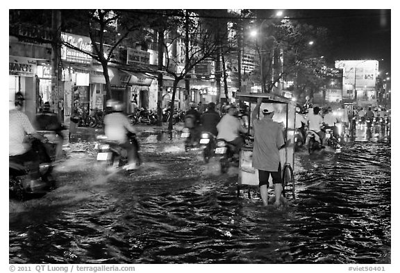 Traffic passes man pushing food cart on flooded street at night. Ho Chi Minh City, Vietnam (black and white)
