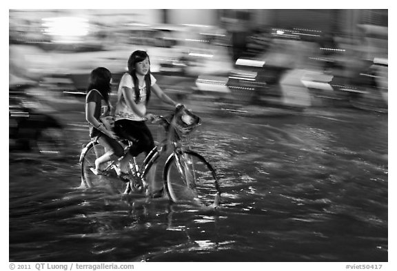 Girls sharing night bicycle ride through water of flooded street. Ho Chi Minh City, Vietnam