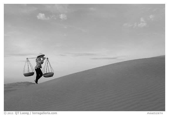 Woman with yoke baskets walks on sand dunes. Mui Ne, Vietnam (black and white)