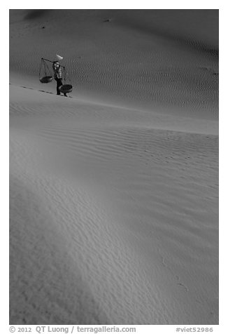 Woman with conical hat and shoulder pole baskets pauses on dune. Mui Ne, Vietnam (black and white)